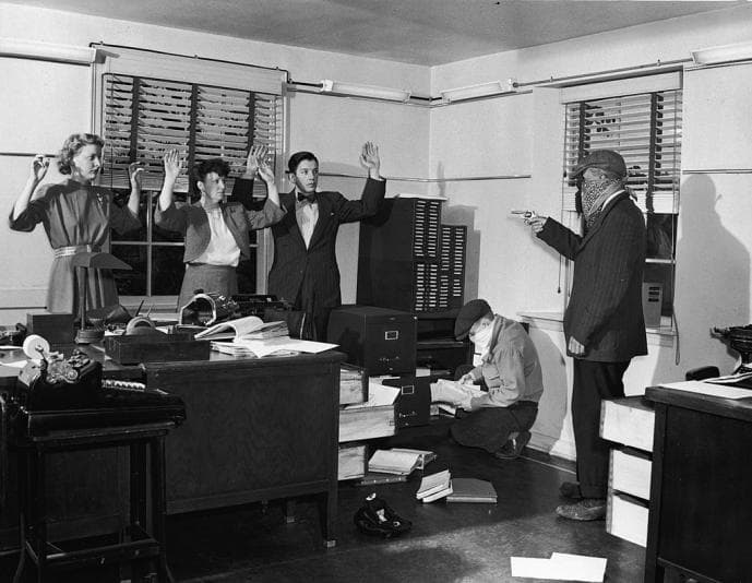 Person with a gun pointing at three people and one person searching drawers