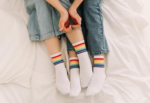 Two pairs of legs, on bed linen, with rainbow pride socks, holding hands