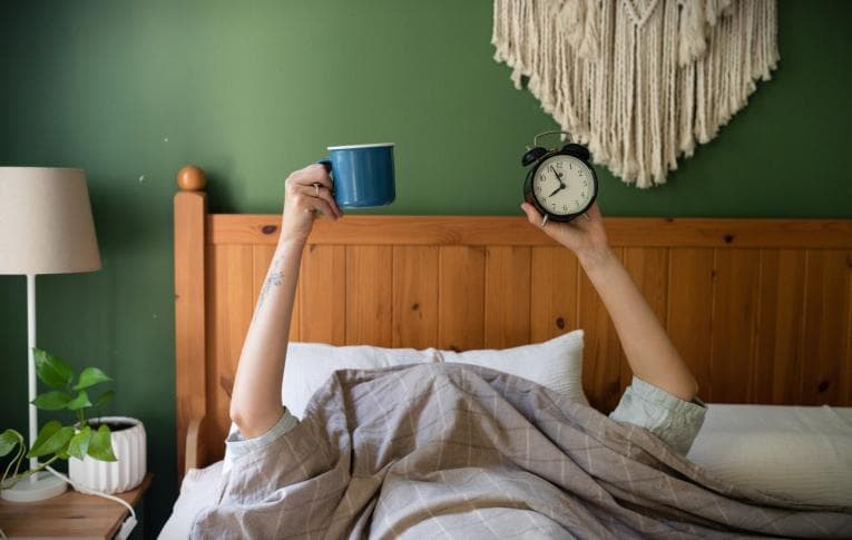 Person in bed holding up a cup of coffee and an alarm clock
